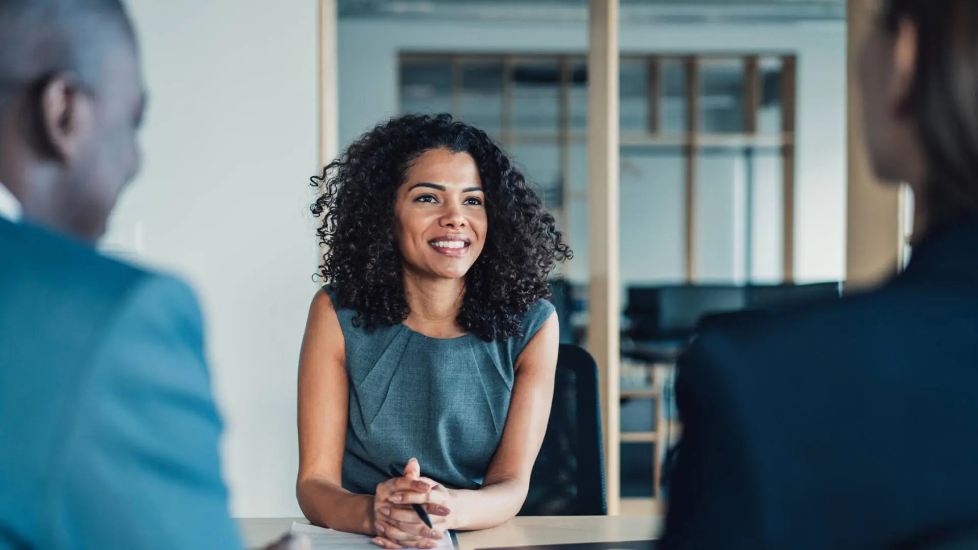 A smiling woman with curly hair is in a meeting, facing two blurred figures in an office.